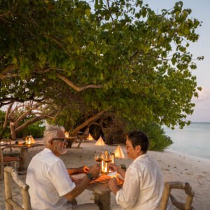 An older couple enjoys a candlelit dinner on the beach at sunset, sitting at a wooden table under leafy trees, with the ocean and additional lit tables in the background.