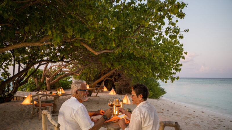 An older couple in white clothing dines at a candlelit table on a sandy beach, surrounded by trees and lanterns, with the ocean and sunset in the background.