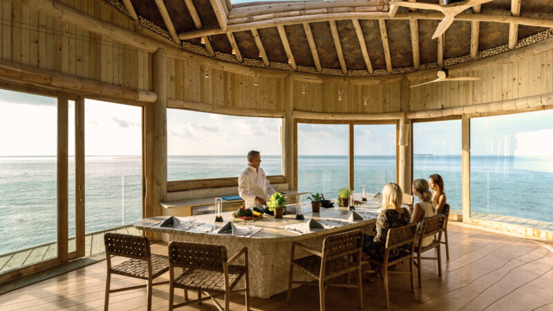 A chef stands at a round dining table set for a meal, talking to three seated people in a sunlit, wooden room with large windows overlooking the ocean.