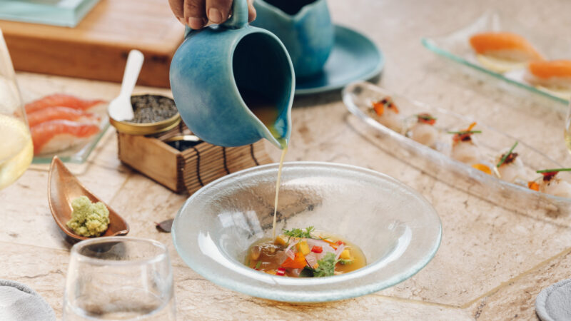 A hand pours golden broth from a blue jug into a glass bowl filled with colorful vegetables and herbs, surrounded by elegant plates of sushi and dining utensils on a stone table.
