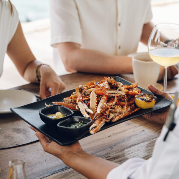 A server presents a platter of crab legs with dipping sauces to two people seated at an outdoor wooden table, with ocean views in the background.