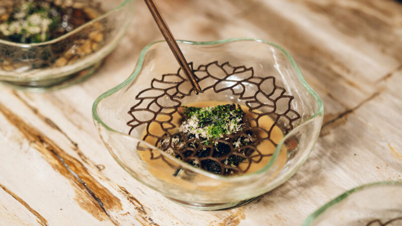 A glass bowl with a decorative, lattice-like garnish on top of a savory dish, garnished with herbs and grated cheese, sits on a rustic wooden surface. Chopsticks are poised over the dish.