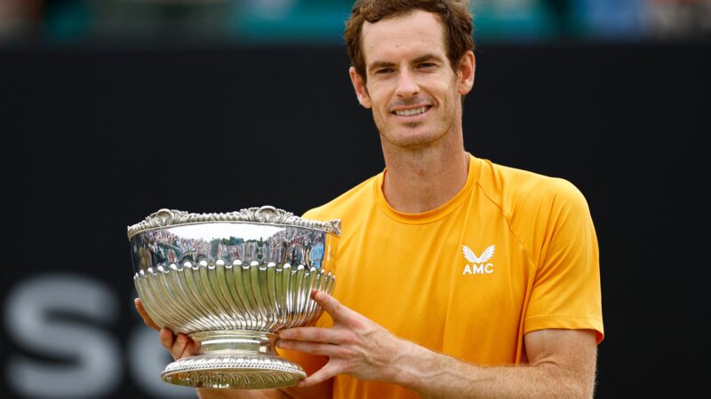 A male tennis player wearing an orange shirt smiles while holding a large silver trophy with both hands on a tennis court, capturing the joy of victory during autumn at Soneva.