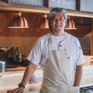 A chef with gray hair, wearing a white shirt and beige apron, stands and smiles in a modern restaurant kitchen with warm lighting and copper hanging lamps.