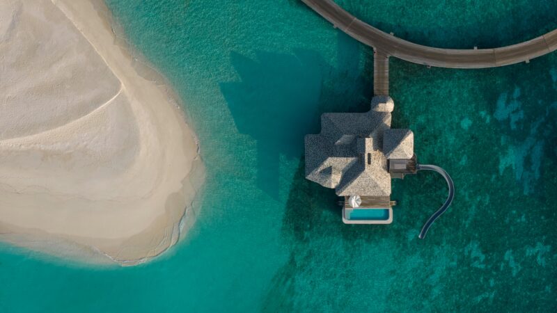 Aerial view of a Soneva Secret villa with a water slide over turquoise ocean water, next to a sandy beach and connected by a wooden walkway.